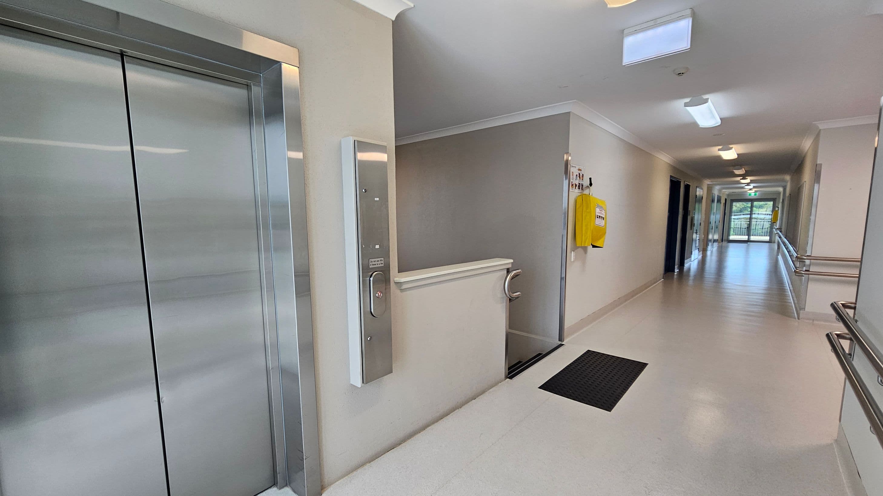 A corridor with a shiny metal lift on the left. Adjacent to the lift is a small platform lift with a control panel. The right side of the corridor features a yellow wall-mounted box and handrails. The floor is light-coloured with a black mat near the platform lift. The ceiling has several rectangular lights, and at the end of the corridor, there is a glass door leading outside.