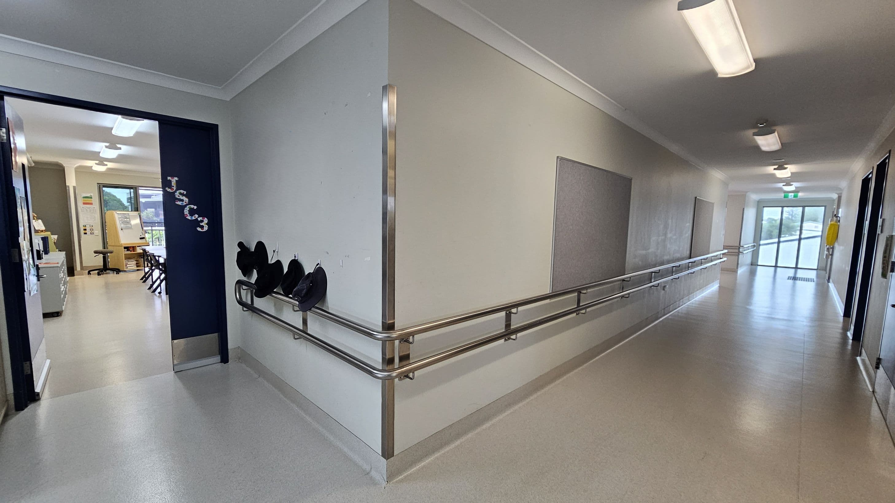 A corridor with a light-coloured floor and ceiling, featuring several overhead lights. On the right, a long handrail runs along the wall, which has a few notice boards. At the end of the corridor, there are glass doors. On the left, an open doorway leads to a classroom with chairs, a desk, and a window. The door has "JSCC3" written on it, and there are hooks with hats hanging on the wall nearby.