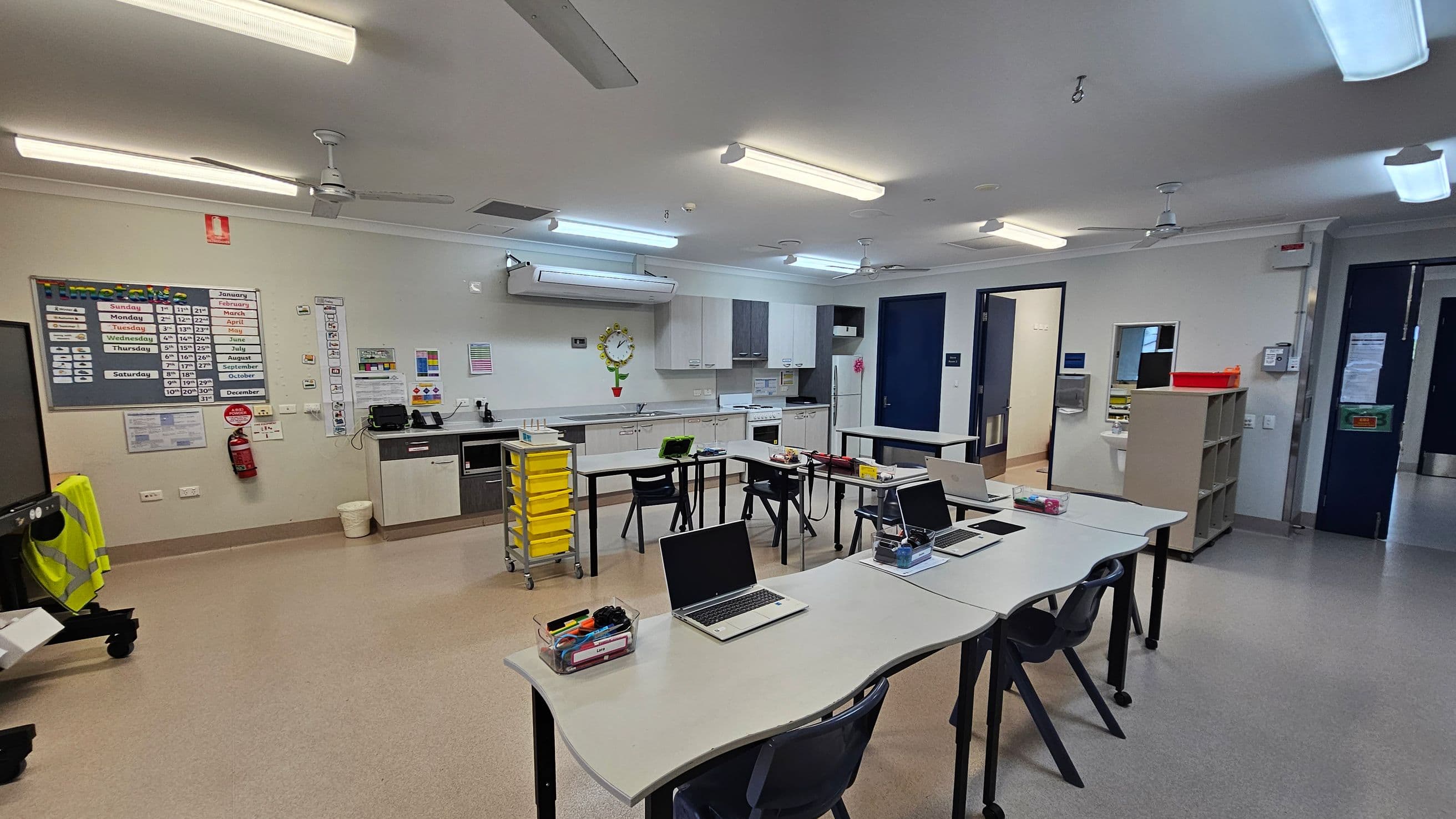 A classroom with a light grey floor and ceiling. In the foreground, there are several white desks with laptops and stationery. The background features a kitchenette with white cabinets, a wall clock, and a yellow storage trolley. On the left wall, there are educational posters and a fire extinguisher. The right side has a doorway and a shelf with a red tray on top. Fluorescent lights and ceiling fans are visible above.