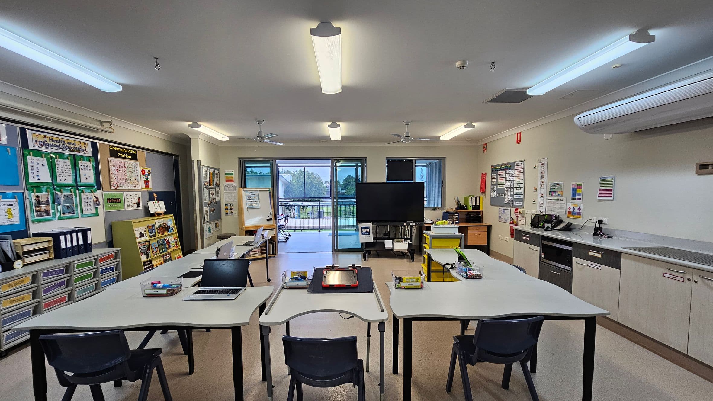 Classroom with a U-shaped arrangement of white tables and black chairs in the foreground. Laptops and stationery are on the tables. The left wall has educational posters and storage drawers. The right side features a counter with cabinets and office equipment. In the centre, a large screen on a stand is near sliding glass doors leading to an outdoor area. The ceiling has fluorescent lights and fans, and the floor is light-coloured.