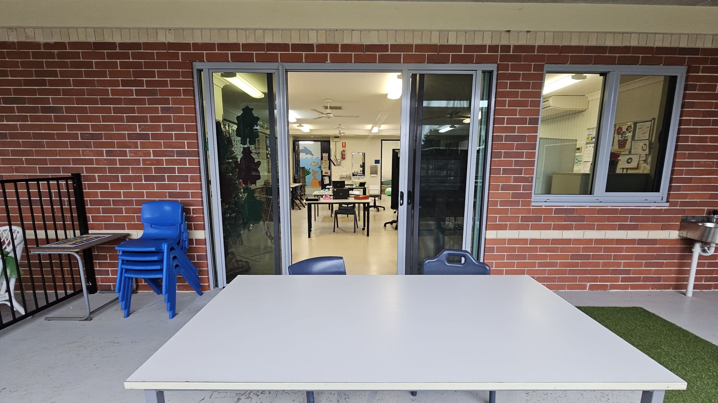 A brick wall with a sliding glass door and a window. In front of the door, there's a white table with a blue chair. To the left, a stack of blue chairs is next to a small table. Inside, a classroom is visible with tables, chairs, and a colourful mural on the far wall. The floor is concrete, and there's a patch of artificial grass on the right. A drinking fountain is mounted on the wall to the right. Ceiling lights are visible inside the room.