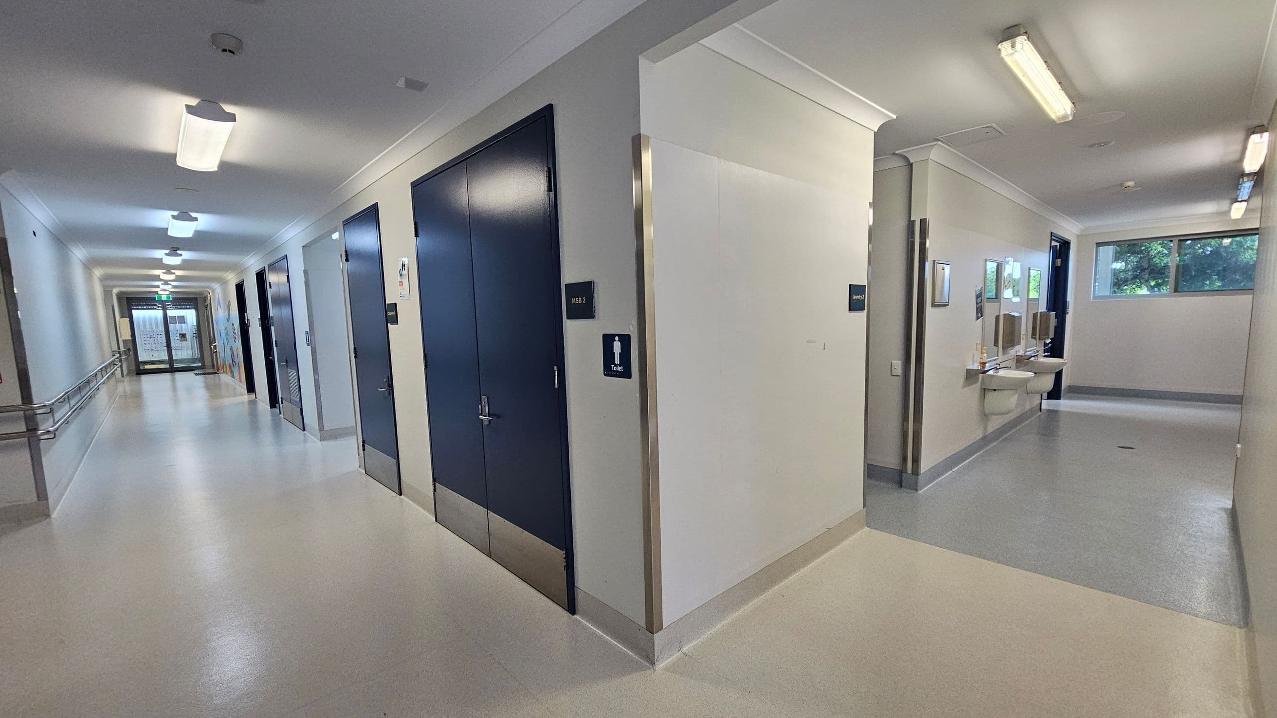 A corridor with a light-coloured floor and ceiling. On the left, there are several doors with signs, and a handrail runs along the wall. On the right, a section leads to a toilet area with sinks and mirrors. Fluorescent lights are mounted on the ceiling. At the end of the corridor, there is a glass door with a view of a window showing greenery outside.