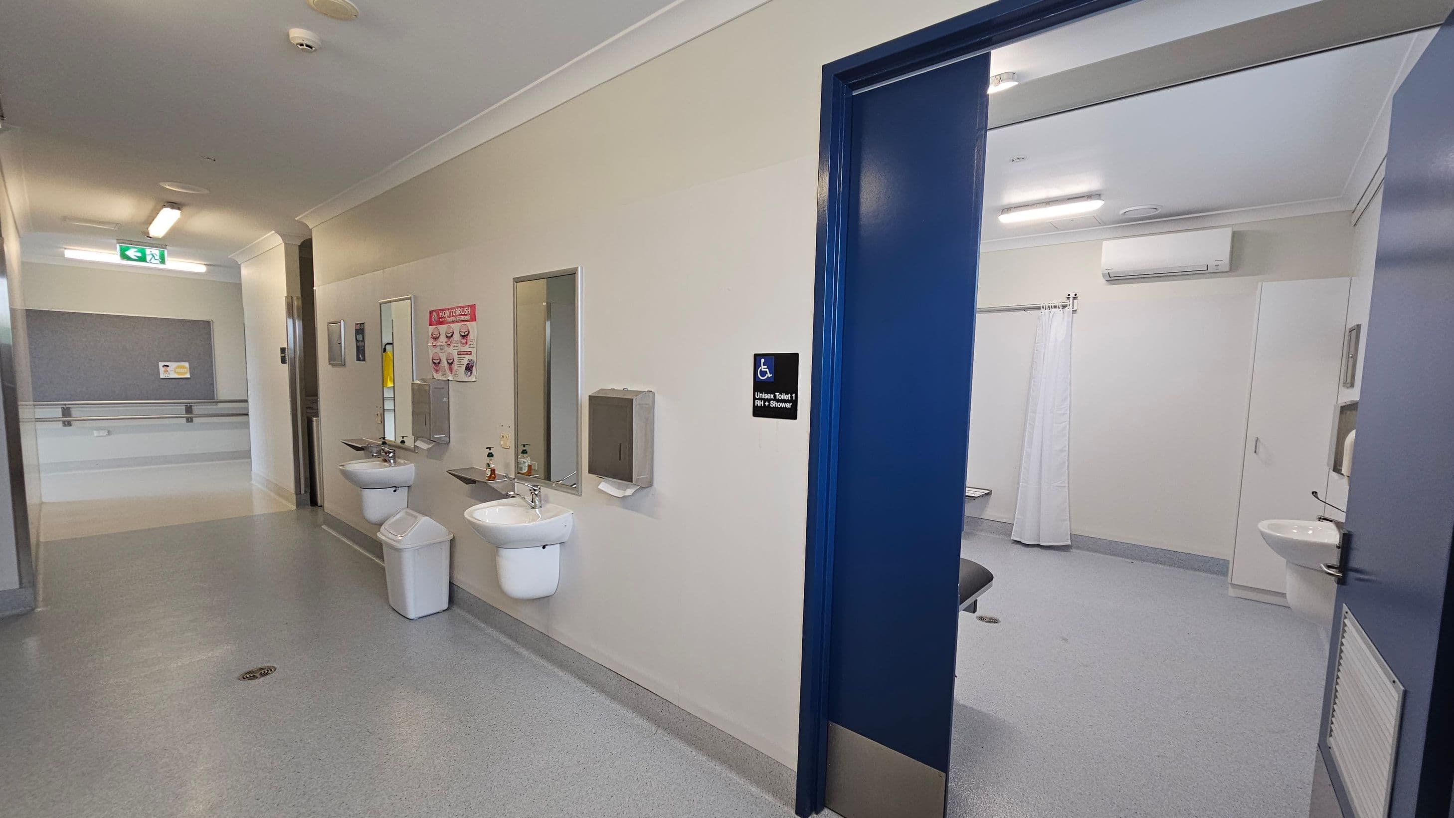 A corridor with a grey floor and white ceiling. On the left, two sinks with mirrors and soap dispensers are mounted on the wall, along with a paper towel dispenser. A sign indicates a unisex toilet with a shower. The right side shows an open door leading to a bathroom with a white curtain and a sink. An air conditioning unit is visible above. The corridor has a handrail and an emergency exit sign.