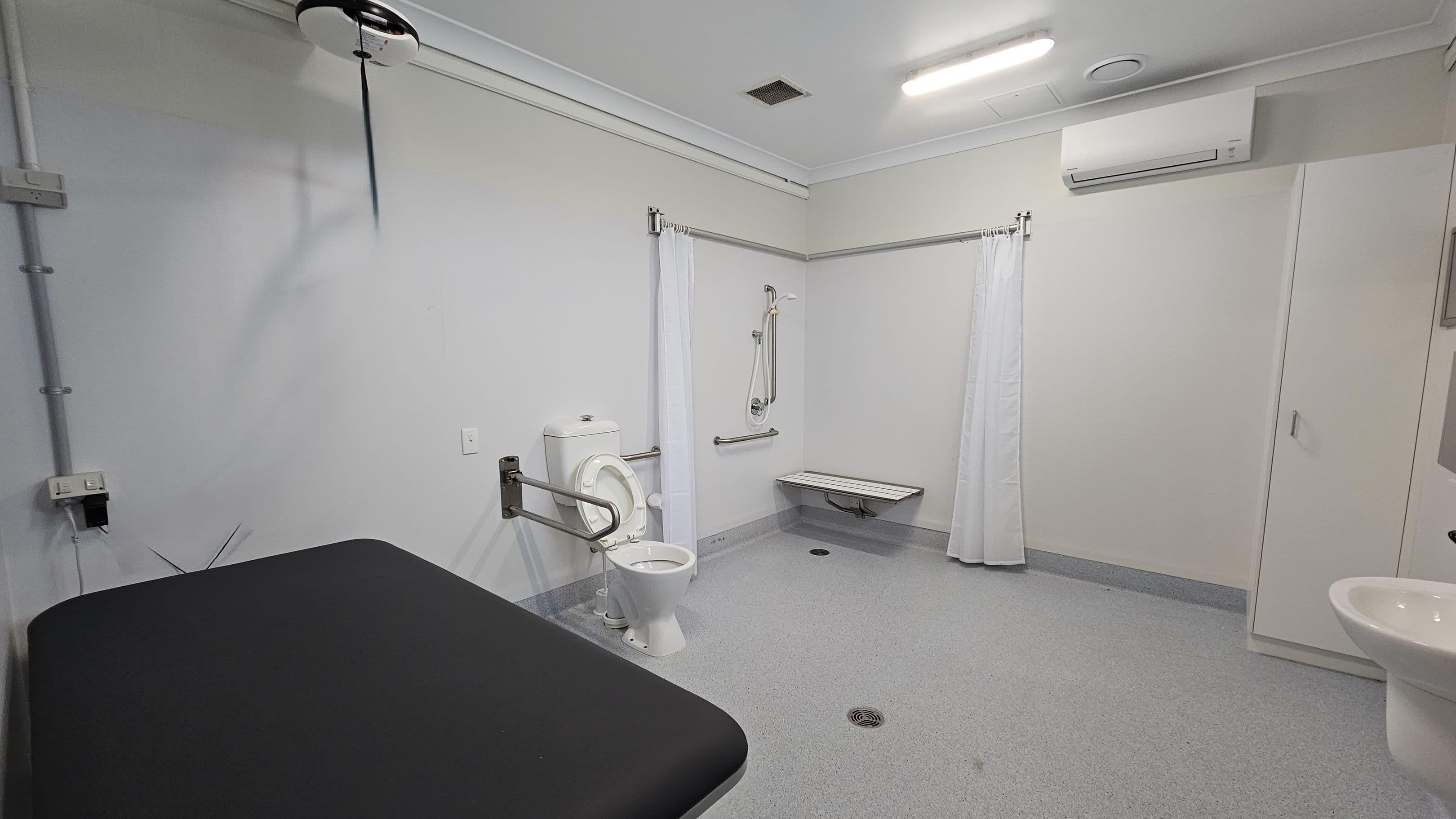A spacious, accessible bathroom with a grey floor and white walls. On the left, a black padded bench is visible. In the centre, a toilet with a support rail is next to a shower area with a foldable seat and a curtain. On the right, a white sink is partially visible, and an air conditioning unit is mounted above a closed cabinet. The ceiling is white with a fluorescent light and a vent.