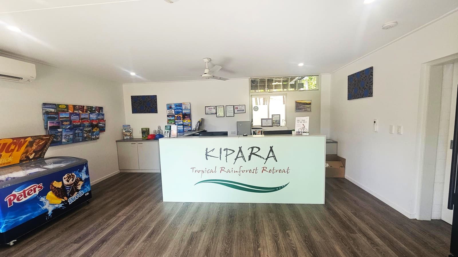 Reception area with a white counter in the centre displaying "KIPARA Tropical Rainforest Retreat". Behind the counter are brochures, certificates, and a window. On the left, a freezer with "Peters" branding and a wall-mounted brochure rack. The floor is wooden, and the ceiling is white with recessed lighting and a ceiling fan.