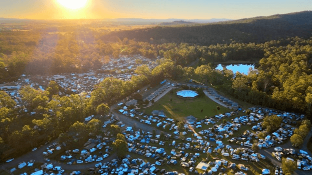 On the left, a dense forest stretches with sunlight filtering through the trees. On the right, a large pond reflects the surrounding greenery. In the middle, a white tent-like structure is surrounded by numerous small tents and vehicles, suggesting a campsite. The background is filled with more trees, creating a lush, green landscape. The foreground features a grassy area with scattered tents and pathways. The floor is grassy, and there is no visible ceiling.