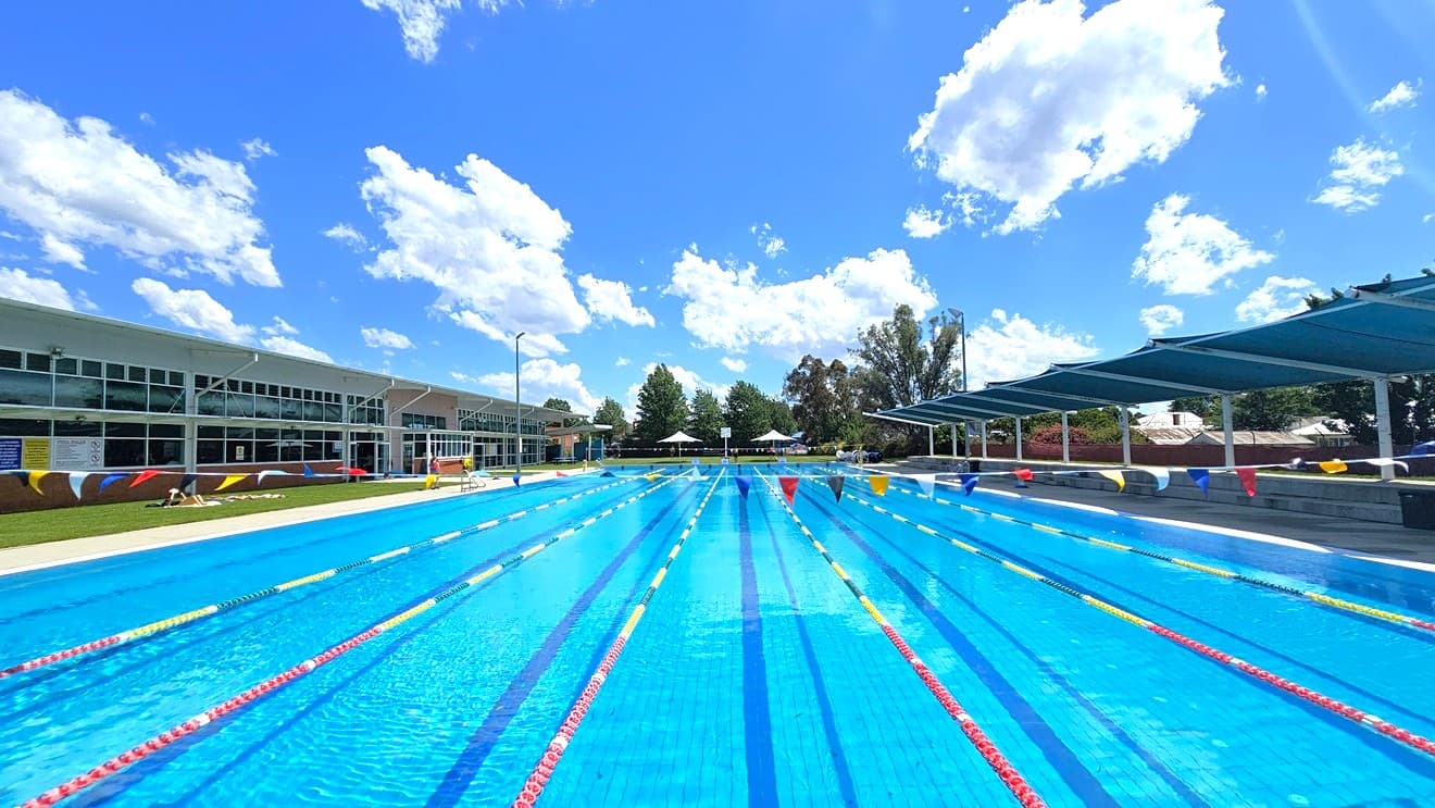 An outdoor swimming pool with multiple lanes, each marked by red and yellow lane dividers. On the left, a modern building with large windows and colourful wall accents. On the right, a series of blue shade structures providing cover. In the background, trees and white umbrellas are visible. The pool is surrounded by a paved area and grass.