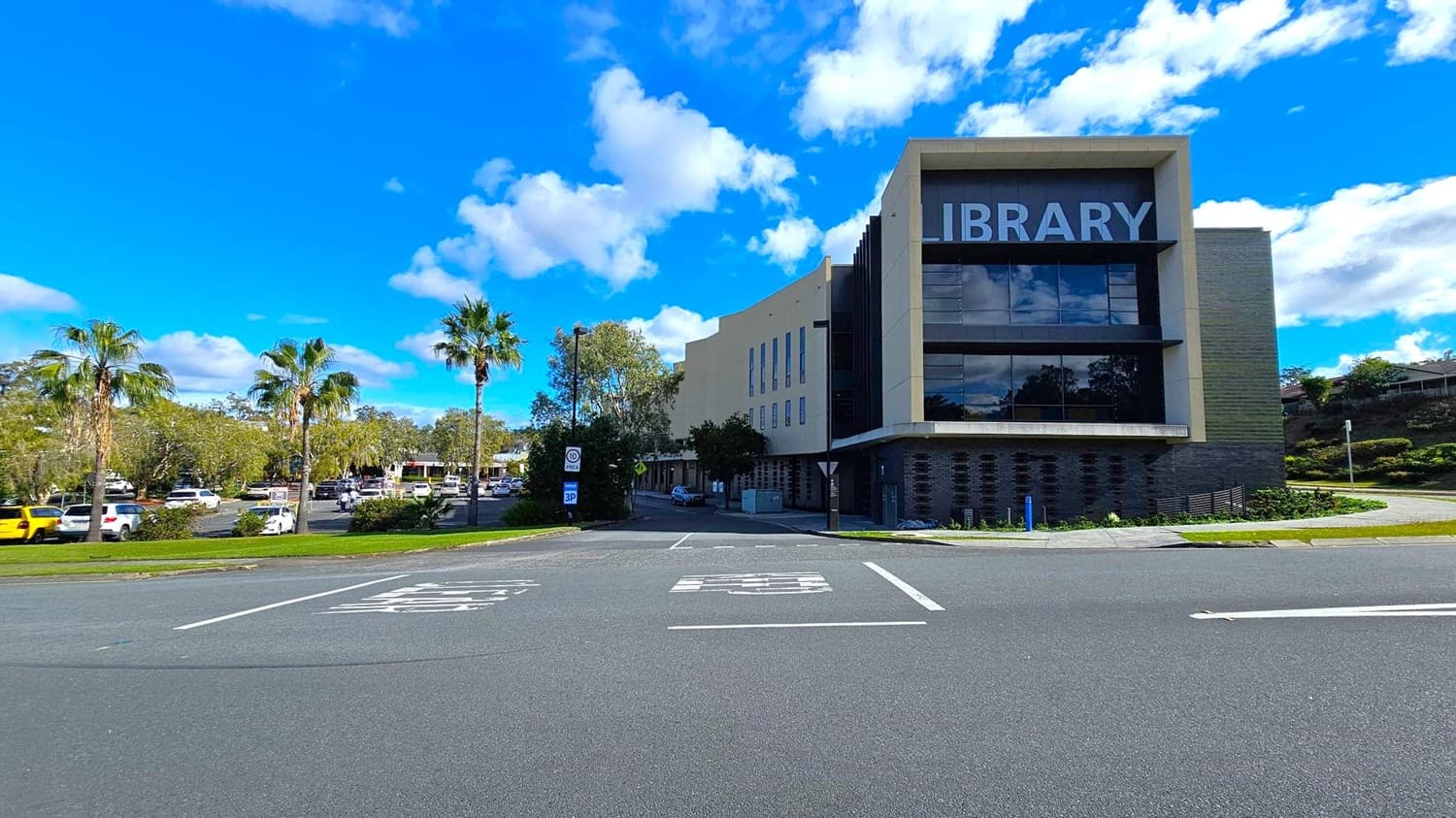 A modern building with the word "BRARY" visible on the right side, likely part of a library sign. The structure is surrounded by palm trees and other greenery. Several cars are parked in a lot to the left, including a yellow one. The foreground features a multi-lane road with white markings. In the background, there are additional buildings and more trees under a blue sky with scattered clouds.