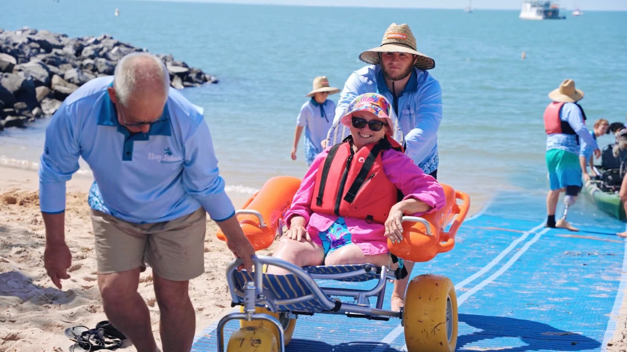 On the left, a beach wheelchair with large yellow wheels and orange armrests sits on grass. On the right, another beach wheelchair with grey wheels is nearby. In the middle, several kayaks and yellow water bikes are lined up on the grass near the shoreline. The background features a calm sea with a few boats and a rocky breakwater. Trees are visible on both sides.