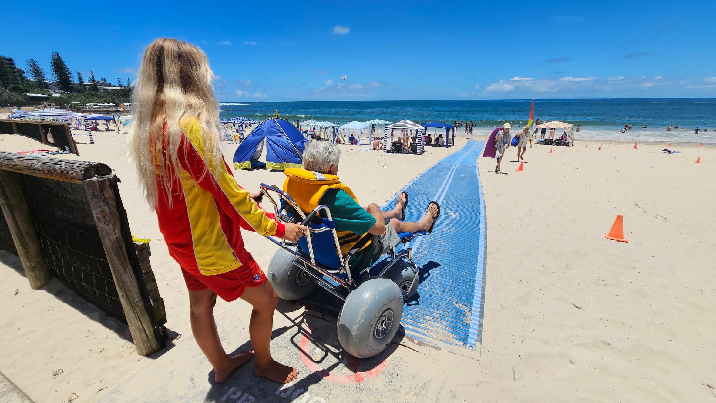 A person with long blonde hair, wearing a red and yellow outfit, is pushing a beach wheelchair with large wheels. The person in the wheelchair is wearing a yellow life jacket and is on a blue mat that leads towards the sea. The beach is busy with people, tents, and umbrellas. Orange cones are placed along the path. The ocean is visible in the background, with people swimming and enjoying the water.