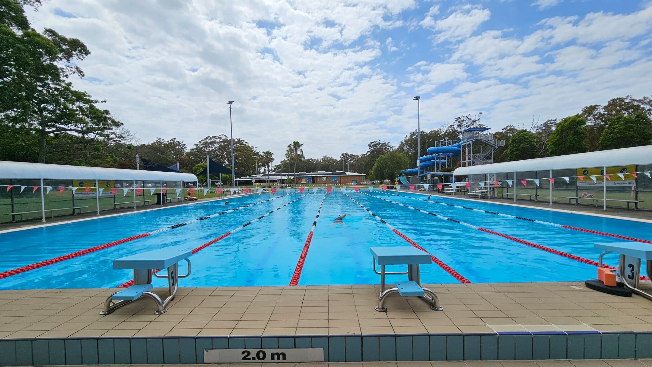 An outdoor swimming pool with clear blue water, marked with red and white lane dividers. Two diving boards are positioned at the pool's edge, with a depth marker indicating 2.0 metres. On the left and right, there are covered seating areas with benches. In the background, a blue water slide structure is visible on the right, surrounded by trees and a grassy area. A few swimmers are in the pool, and a building is seen in the distance.