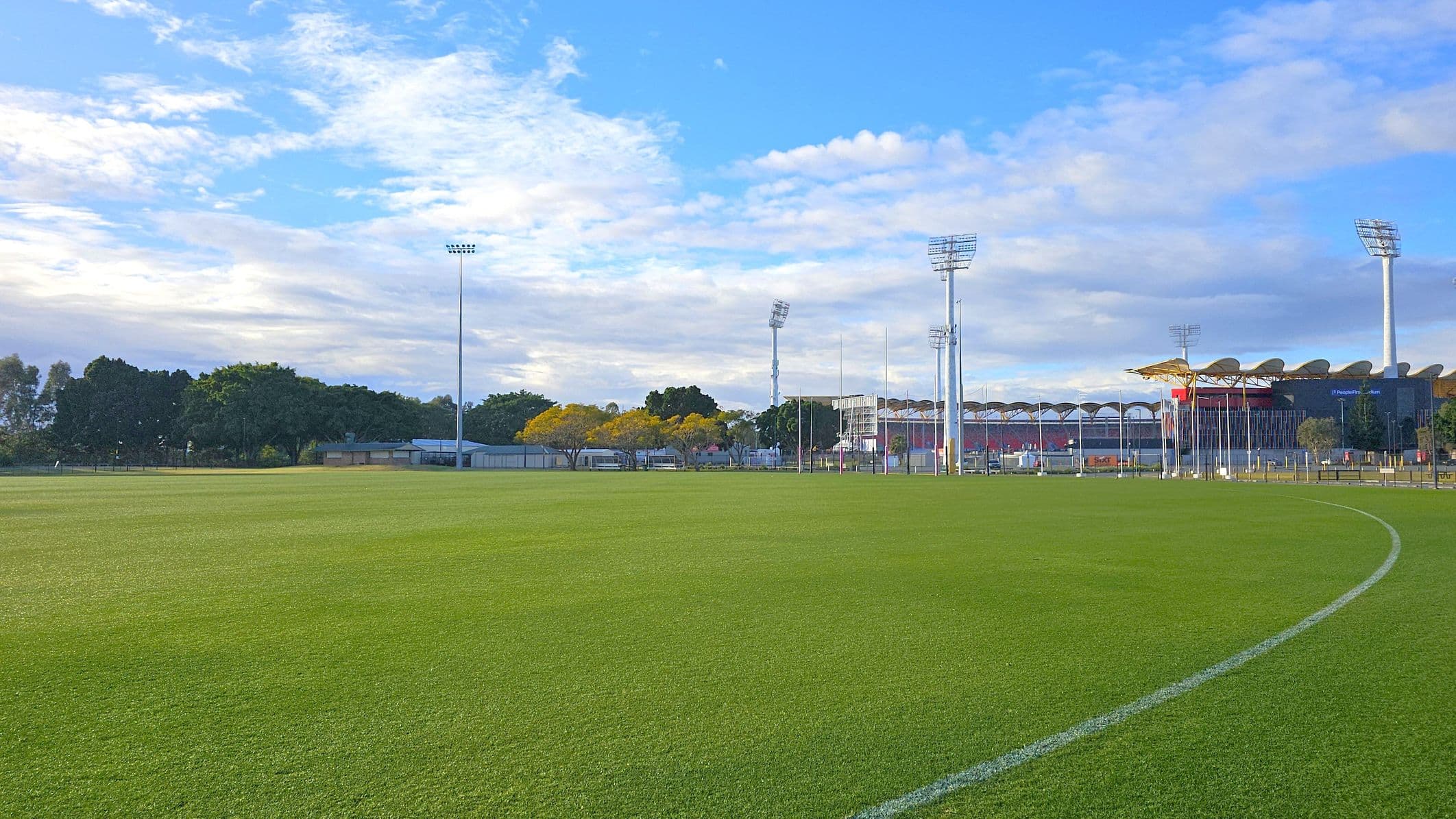 A large, open sports field with well-maintained grass. In the centre, there are goalposts in the distance. The foreground shows a curved white line on the grass. To the right, a tall floodlight pole stands near the edge of the field. The background features a line of trees and some buildings, with a few parked cars visible. Red signs with "GOLDCOAST" are placed along the perimeter fence.