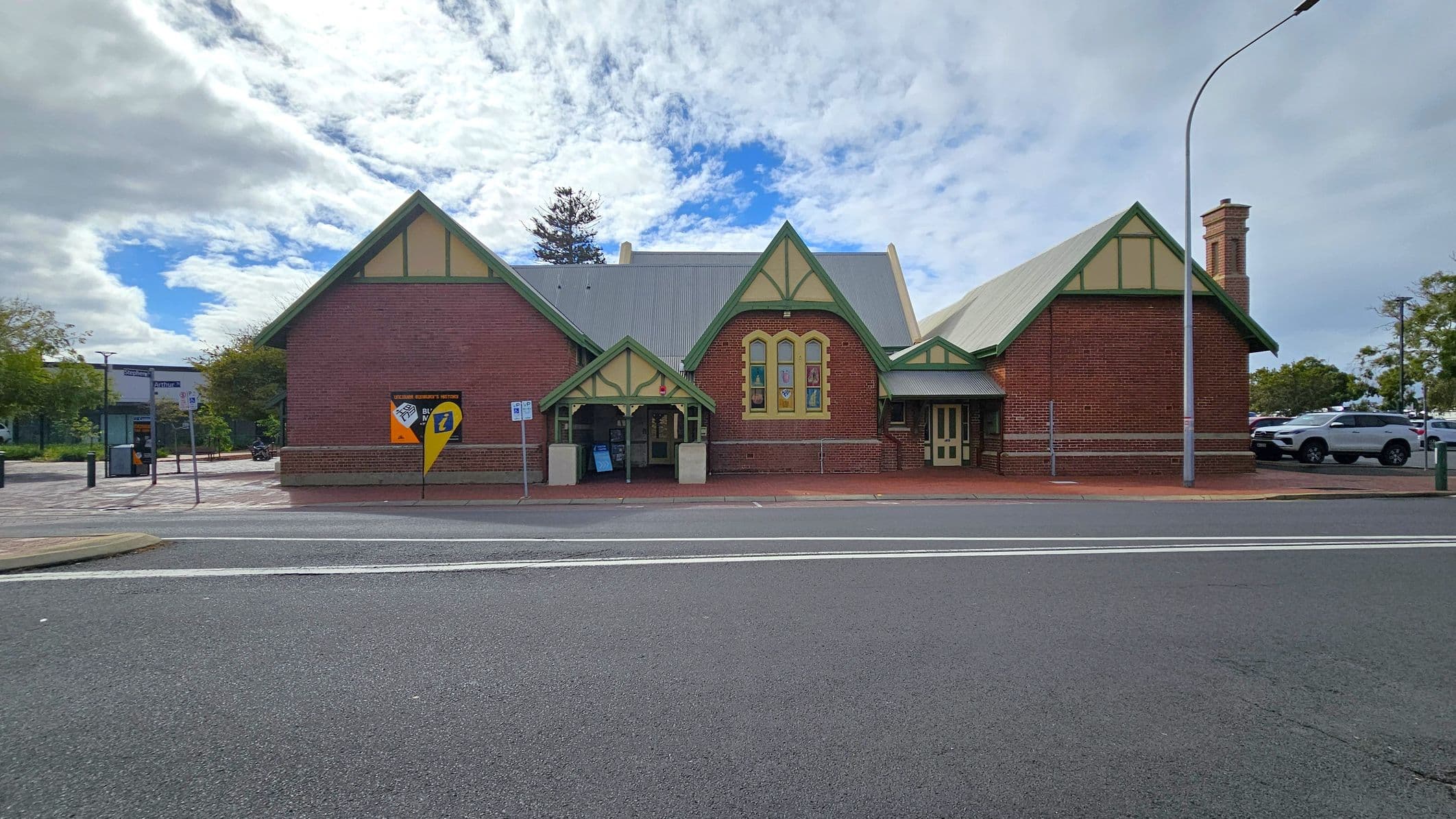 A historic brick building with a gabled roof and green trim stands prominently. It features arched windows and a small entrance with a canopy. A yellow and blue sign is displayed on the left side. The foreground shows a paved road with white lines, and a footpath runs along the building. A streetlamp is positioned to the right, and there are parked cars and trees in the background.