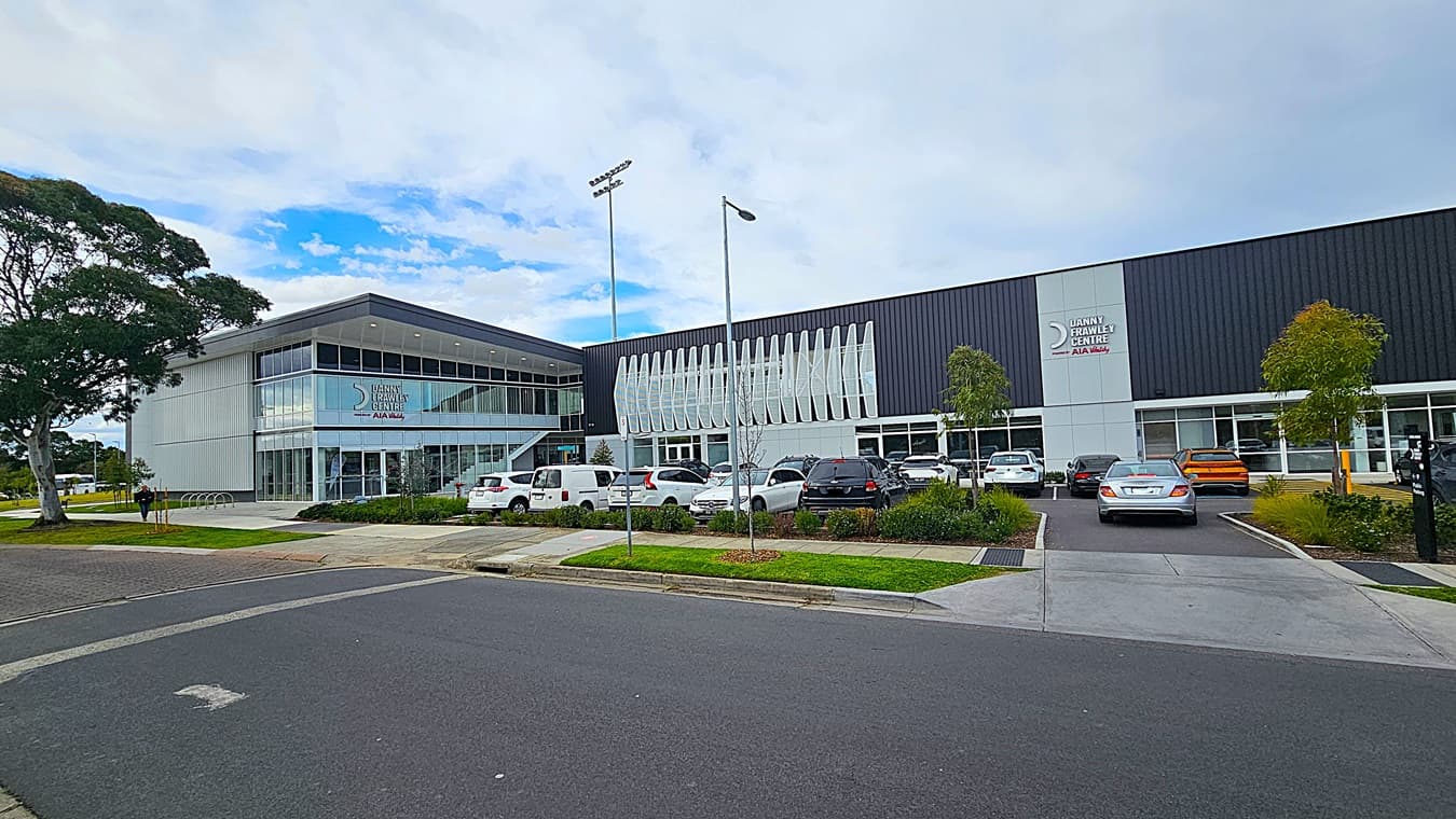 A modern building with a large glass facade on the left, featuring the "Danny Frawley Centre" sign. The right side has a black and white exterior with vertical louvres. In the foreground, there is a car park with several vehicles, including white, black, and orange cars. The background includes a few trees and a well-maintained lawn.