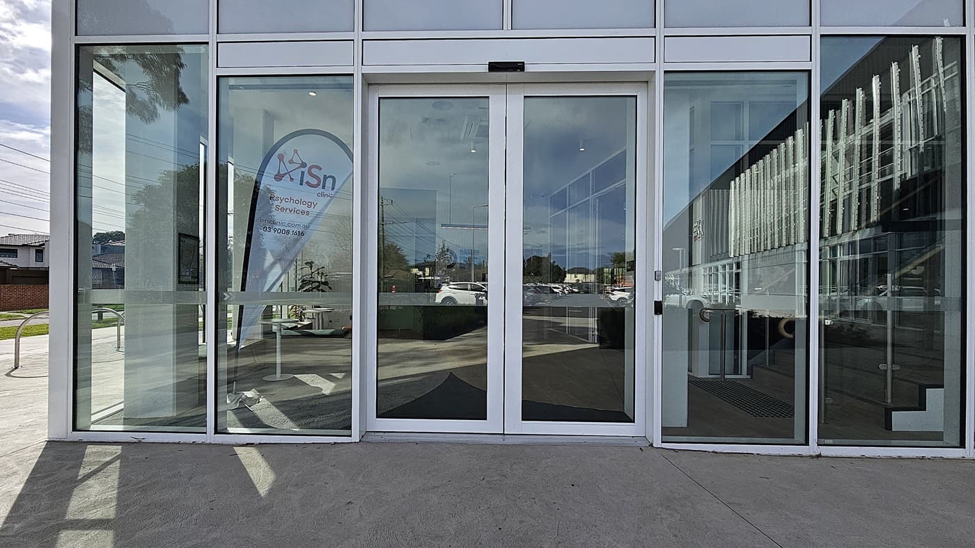On the left, a green door with open shelving beside it, and a reception desk with a dark green marble front. In the middle, a white wall with the "Danny Frawley Centre" logo and a wall-mounted television. On the right, glass doors leading to a gym area and a wooden staircase with a metal handrail. The floor is light wood, and the ceiling is white with recessed lighting and a black linear light fixture.