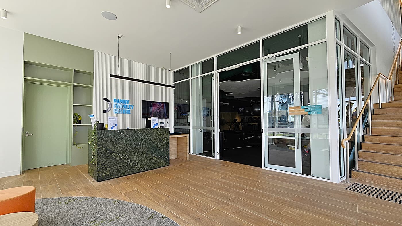 On the left, a wall-mounted television above a wooden desk with a blue device on it. In the middle, double glass doors open to a gym area with exercise equipment and green ceiling panels. On the right, a staircase with wooden steps and a glass railing leading to an upper level. The floor is light wood, and the ceiling is white with recessed lighting and a linear light fixture.
