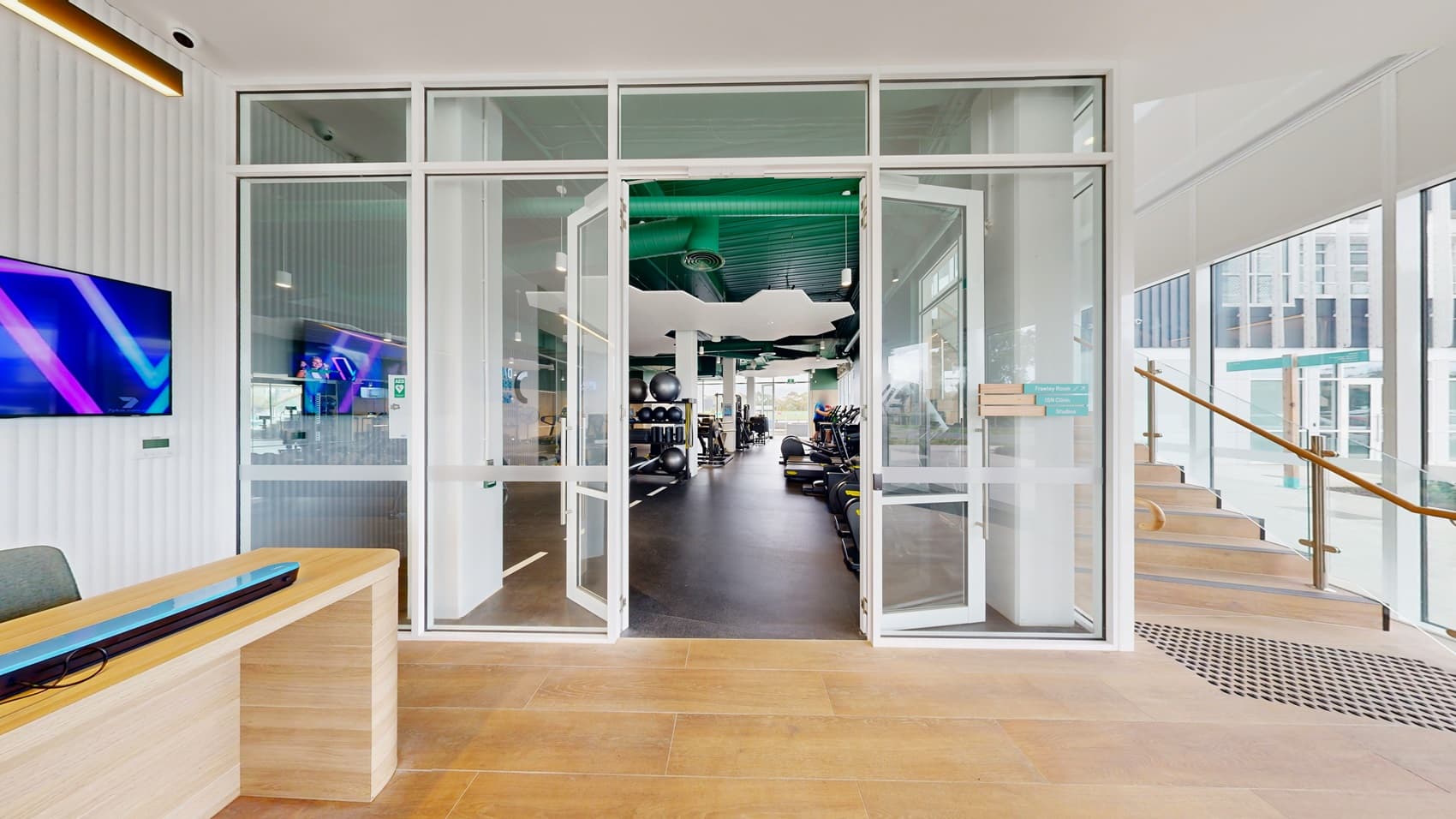 On the left, a long rack of dumbbells and weightlifting equipment. In the middle, various exercise machines, including treadmills and rowing machines, with a white pillar and a wall-mounted television. On the right, shelves holding large exercise balls and foam rollers. The floor is dark rubber, and the ceiling is green with white, cloud-shaped acoustic panels and hanging light fixtures.