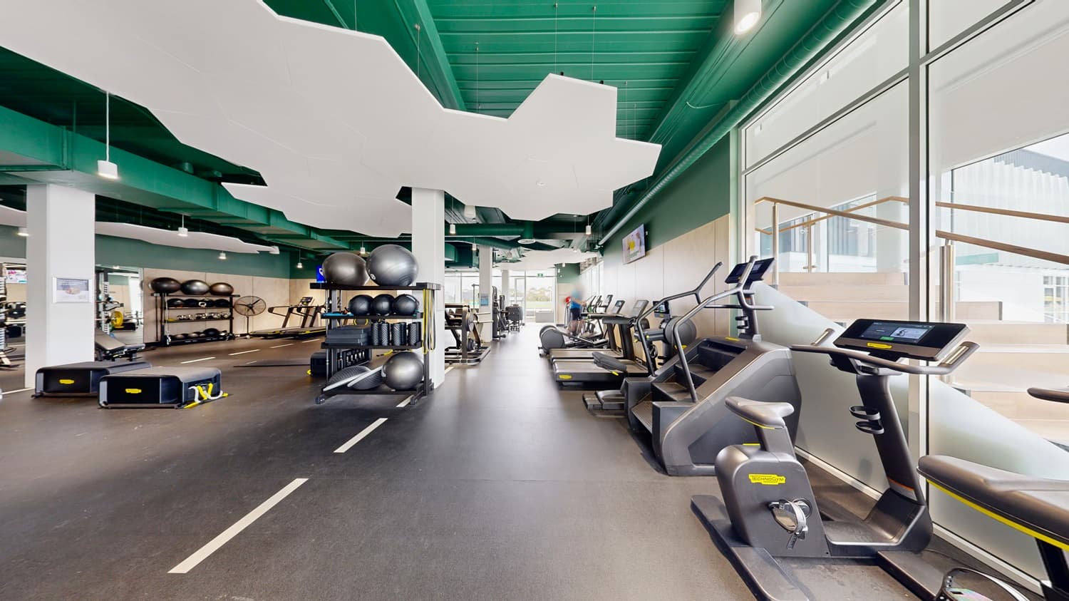 On the left, a white pillar next to shelves holding large exercise balls and balance boards. In the middle, various exercise machines, including treadmills and ellipticals, with a white pillar and a wall-mounted television. On the right, a staircase with wooden steps and a glass railing leading to an upper level. The floor is dark rubber, and the ceiling is green with white, cloud-shaped acoustic panels and hanging light fixtures.