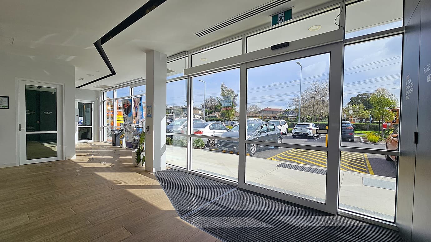 On the left, a white door with a glass panel and a black sign next to it. On the right, large glass sliding doors with a green exit sign above. In the middle, a white column and a potted plant. The background includes a car park with several vehicles and a row of houses. The foreground has a wooden floor with a black mat near the entrance. The ceiling is white with black recessed lighting and ventilation panels.