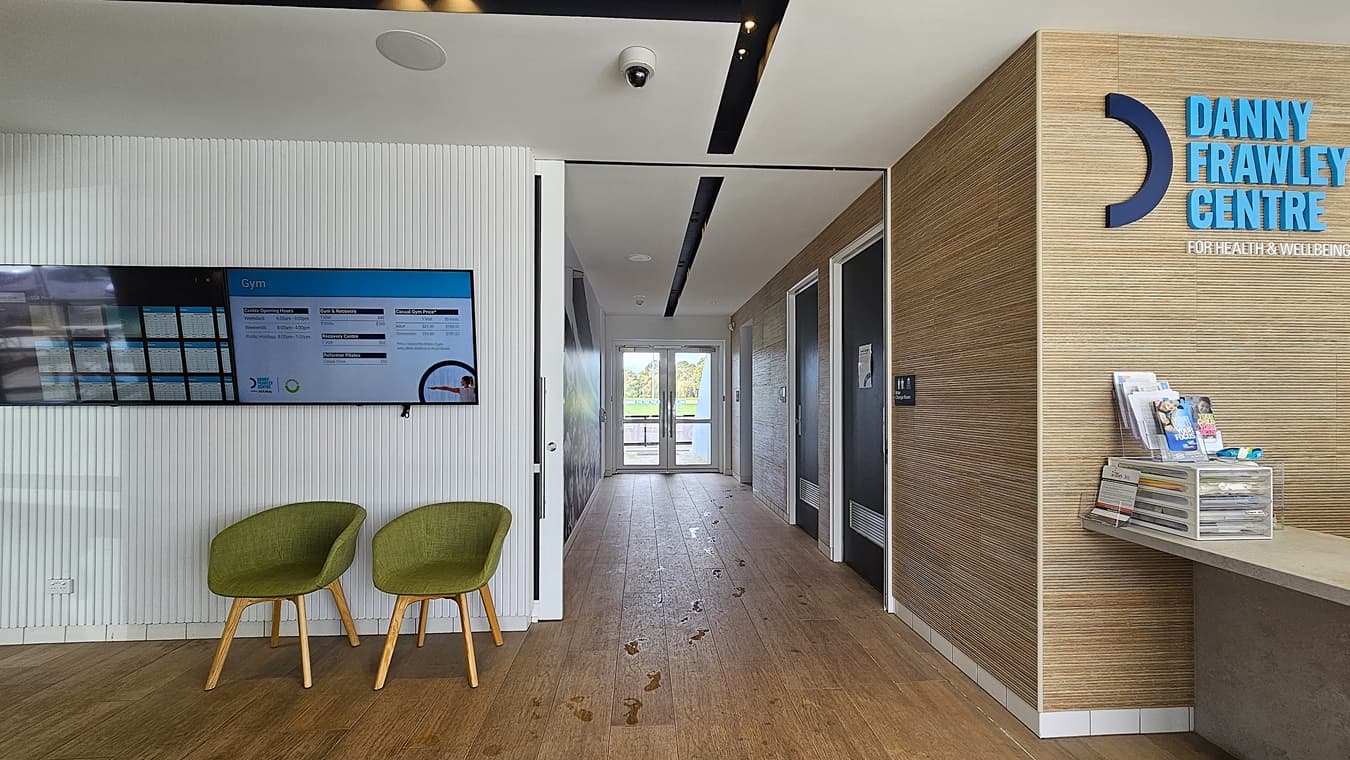 On the left, two green chairs are placed beneath a wall-mounted screen displaying information. On the right, a wooden panel wall with the "Danny Frawley Centre" sign and a reception counter holding brochures and a clear donation box. In the middle, a hallway with wooden flooring leads to glass double doors, revealing greenery outside. The floor is wooden with visible footprints, and the ceiling is white with black recessed lighting and a security camera.