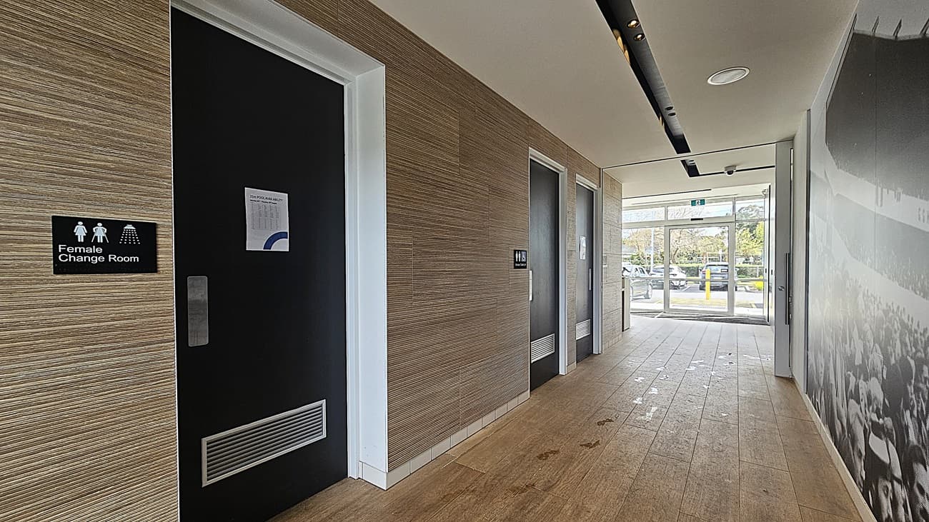 On the left, a black door labelled "Female Change Room" with a white frame and a wood-textured wall. On the right, a large black and white mural of a crowd. In the middle, a hallway with three black doors and a glass exit door at the end, revealing parked cars and greenery outside. The floor is light brown tile with visible footprints, and the ceiling is white with black recessed lighting and ventilation panels.