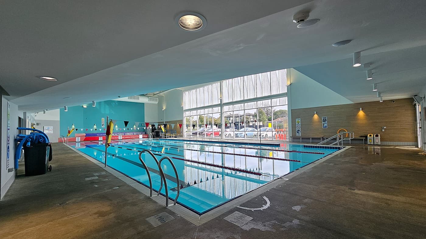 On the left, a black bin with blue pool cleaning equipment and a white wall with safety signs. On the right, a wooden panel wall with posters, two yellow and white bins, and a small bench. In the middle, an indoor swimming pool with lane dividers and metal handrails. The background features large windows showing parked cars and trees. The floor is concrete with wet patches, and the ceiling is white with recessed lighting and exposed ducts.