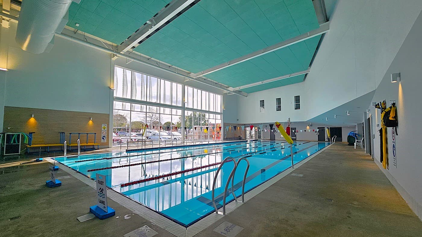 On the left, a wooden panel wall with mounted lights and poolside equipment, including blue and green pool noodles. On the right, a white wall with hanging life jackets and a concrete walkway. In the middle, an indoor swimming pool with lane dividers and metal handrails. The background features large floor-to-ceiling windows showing parked cars and trees. The floor is concrete with wet patches, and the ceiling is turquoise with white beams and exposed ducts.