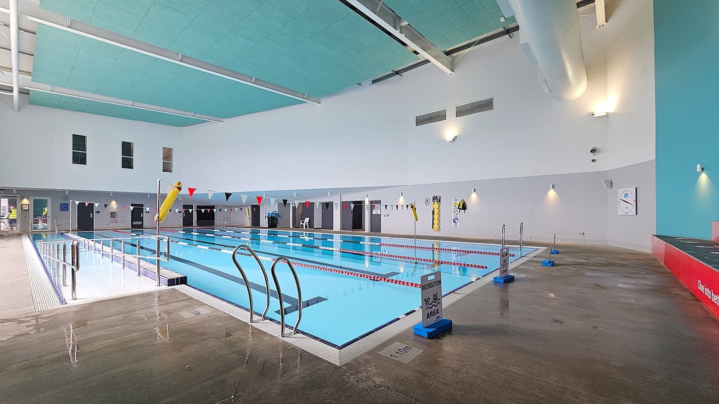 On the left, a shallow pool area with metal handrails and a wet concrete floor. On the right, a red platform with a "Life Time" sign and a deeper pool section. In the middle, a large indoor swimming pool with lane dividers and colourful triangular flags overhead. The background features a white wall with safety equipment and doors leading to changing rooms. The ceiling is turquoise with white beams and large ventilation ducts.