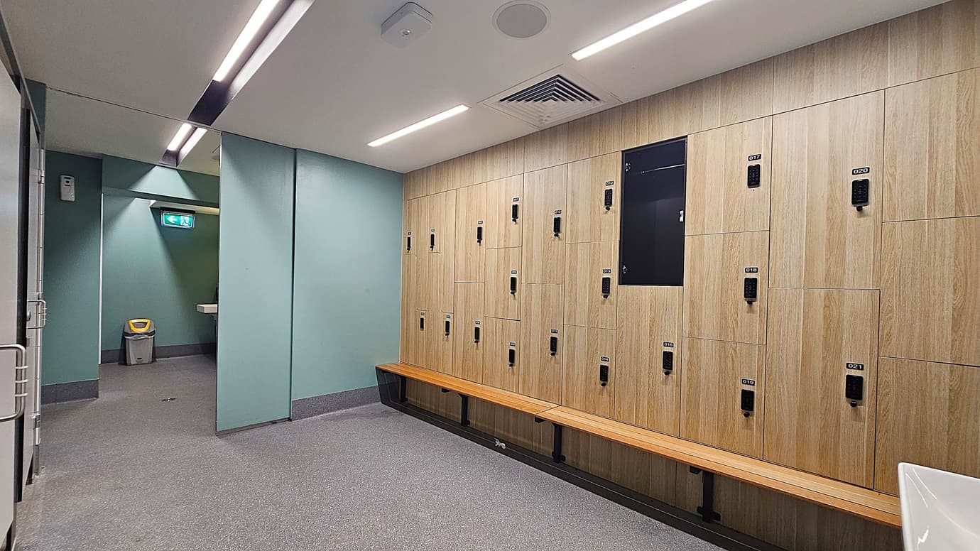 On the left, a green wall with a grey bin and a white sink partially visible. On the right, a wooden panel wall with multiple lockers and a long wooden bench beneath them. In the middle, a grey carpeted floor leading to a hallway with another green wall and an emergency exit sign. The ceiling is white with recessed lighting and ventilation panels.