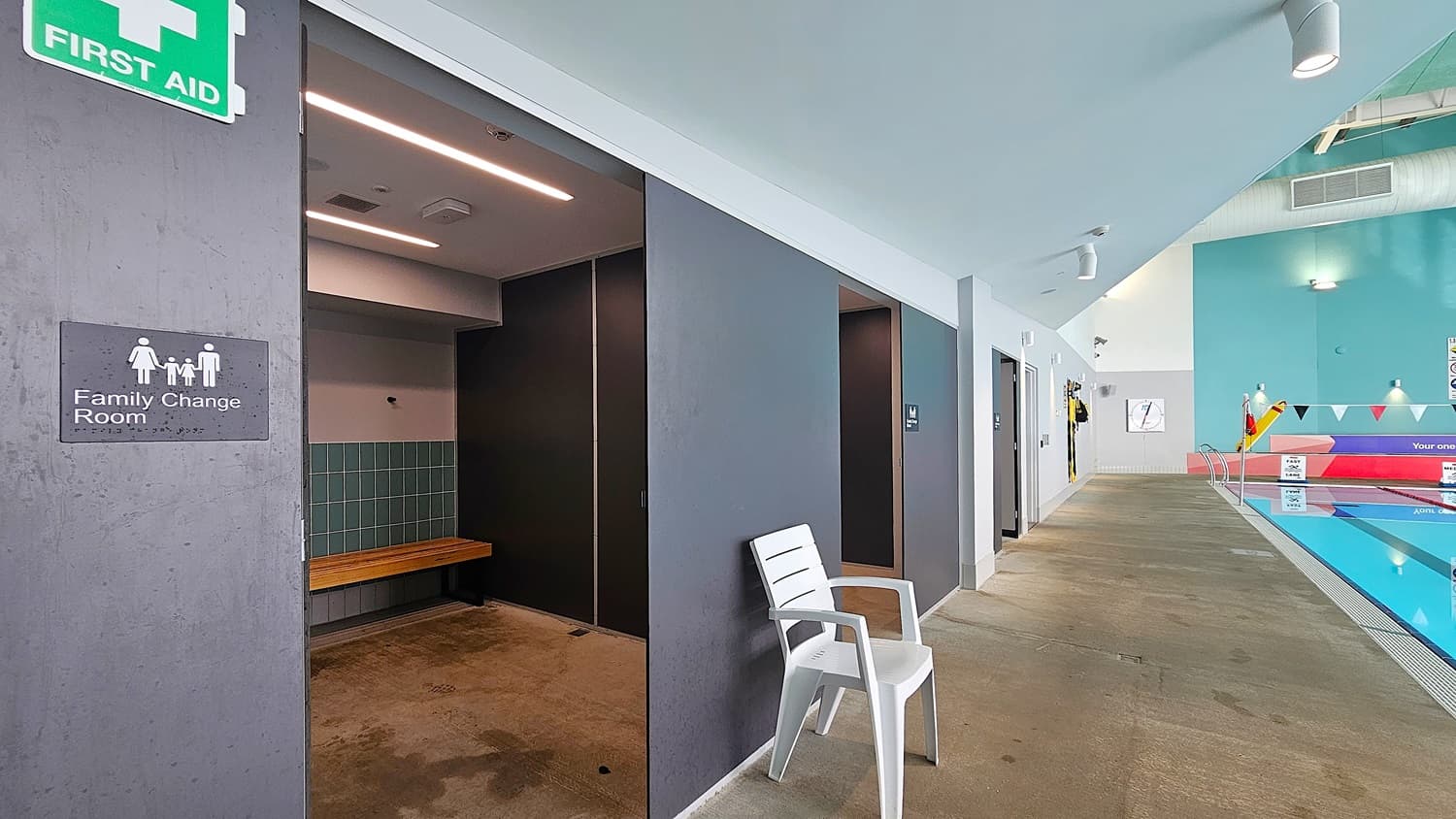 On the left, a grey wall with a "Family Change Room" sign and a green "First Aid" sign above. On the right, a white plastic chair against a grey wall with doors leading to changing rooms. In the middle, a corridor with a concrete floor leading to a swimming pool area. The background features a turquoise wall with safety equipment and a pool with lane dividers. The ceiling is white with recessed lighting and exposed ducts.