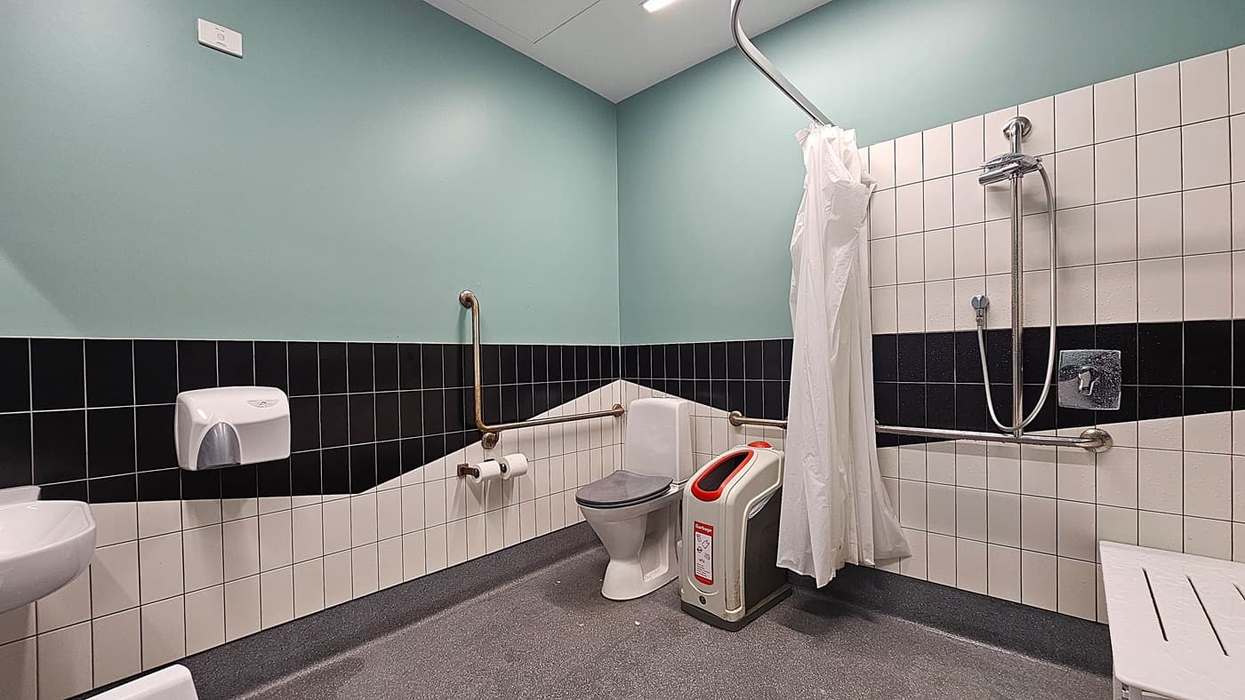 On the left, a light green wall with a white paper towel dispenser and a metal grab bar above a toilet paper holder. On the right, a white tiled wall with a black stripe, featuring a showerhead, a metal grab bar, and a white shower curtain. In the middle, a white toilet with a grey seat, a red and white bin, and a white shower chair. The floor is grey with a textured surface, and the ceiling is white with recessed lighting.