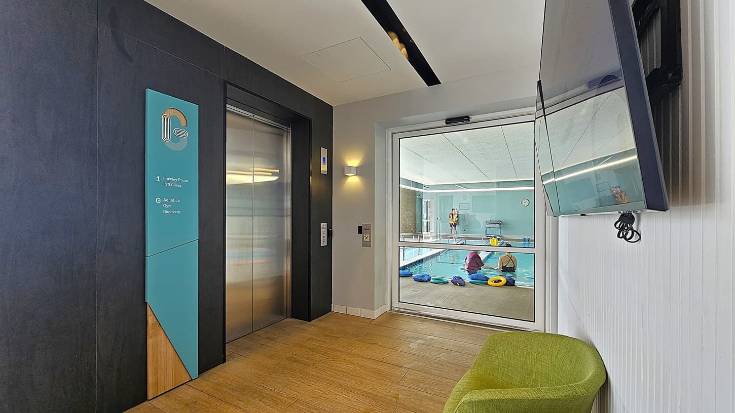 On the left, there is a metallic lift with a blue and wooden sign next to it, indicating different floors. In the middle, a glass door reveals an indoor swimming pool area with people and colourful pool floats. On the right, a large wall-mounted television screen is visible above a green chair. The floor is wooden, and the ceiling is white with a black strip light.