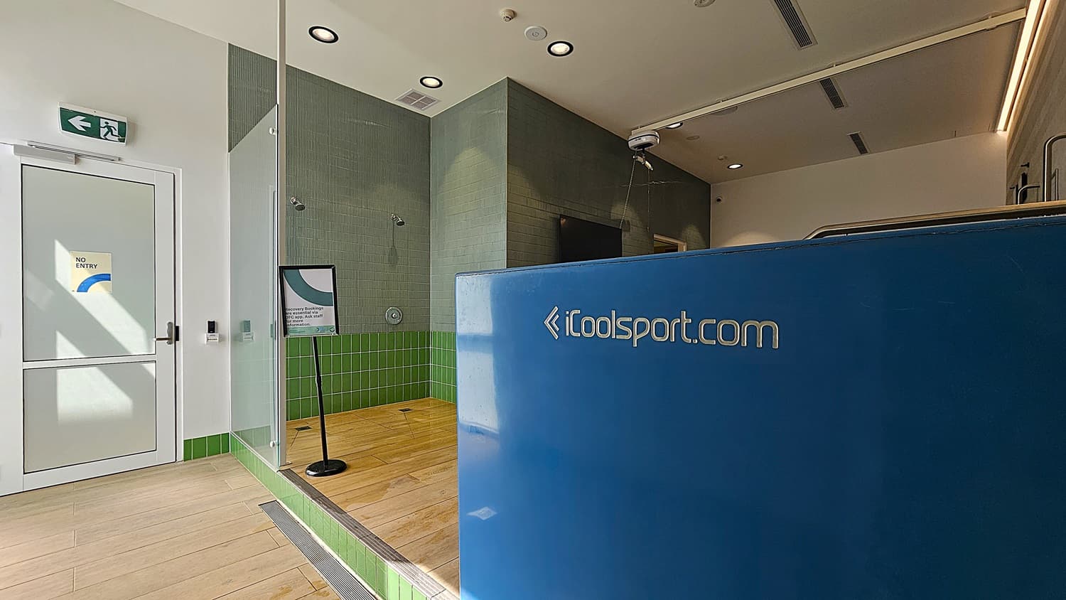 On the left, a white door with a "No Entry" sign and a green exit sign above it. In the middle, a glass partition separates the shower area with green tiles and a sign on a stand. On the right, a large blue cold plunge pool with the text "iCoolsport.com" on it. The floor is light wood, and the ceiling is white with recessed lighting and ventilation panels.