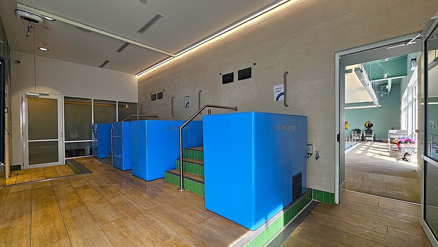 On the left, a white door with a glass panel and a metal handle. In the middle, three blue cold plunge pools with green-tiled steps leading up to them, each with a metal handrail. On the right, an open glass door leads to a brightly lit room with a swimming pool and various equipment. The floor is light brown tile, and the ceiling is white with recessed lighting and ventilation panels.