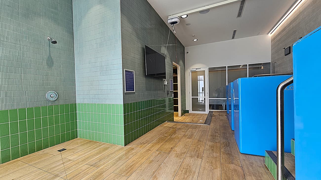 On the left, a green-tiled wall with a showerhead and a drain on the floor. In the middle, a wall-mounted television above a small notice board, with a doorway leading to another room. On the right, three blue cold plunge pools with metal handrails and green-tiled steps. The floor is light wood, and the ceiling is white with recessed lighting and ventilation panels.