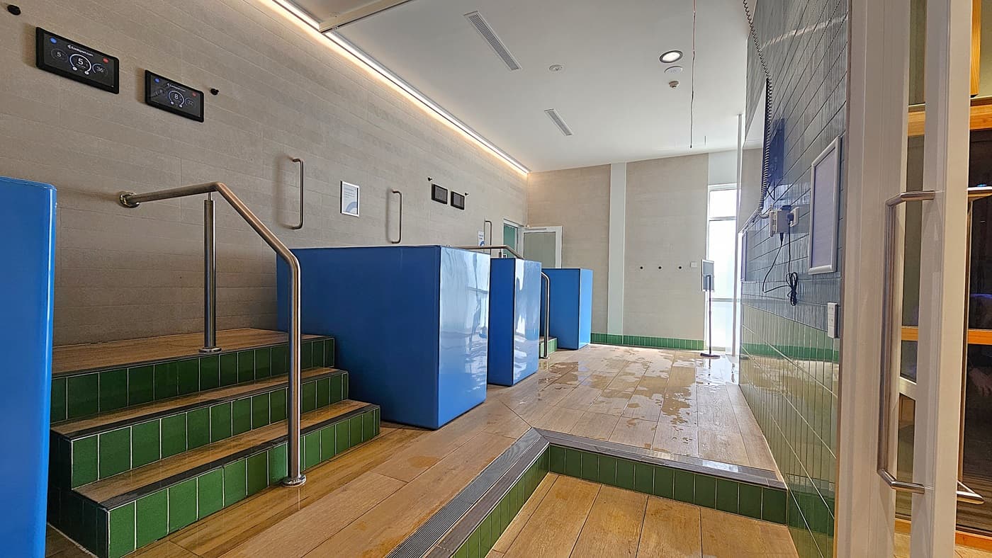 On the left, green-tiled steps with a metal handrail lead up to three blue cold plunge pools. In the middle, a light-coloured wall with control panels and handrails, and a doorway with a glass door. On the right, a green-tiled wall with a wall-mounted television and a notice board. The floor is light wood with some water puddles, and the ceiling is white with recessed lighting and ventilation panels.