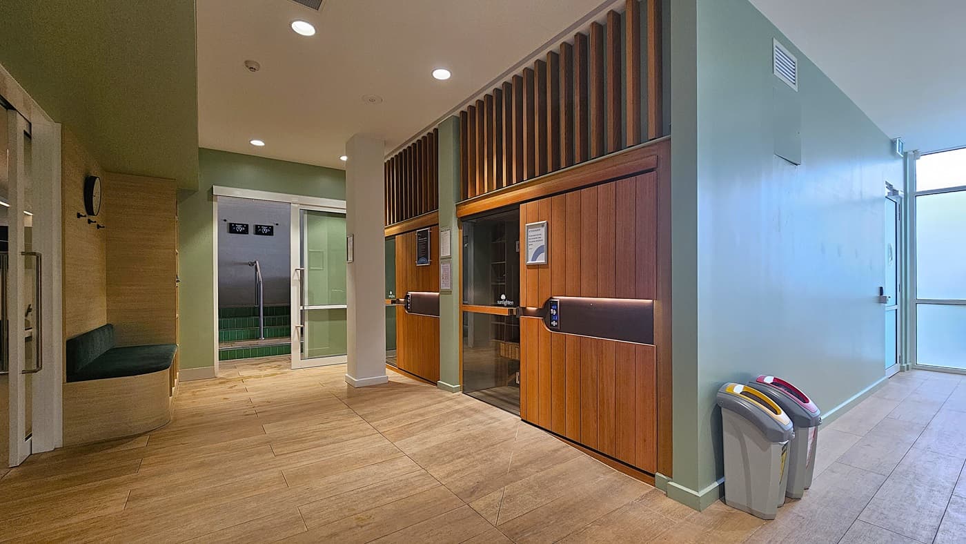 On the left, a wooden bench with a green cushion beneath a wall-mounted clock. In the middle, a glass door leads to a room with green-tiled steps and a metal handrail. On the right, two wooden sauna doors with control panels and signs, and two waste bins with coloured lids. The floor is light wood, and the ceiling is white with recessed lighting and ventilation panels.