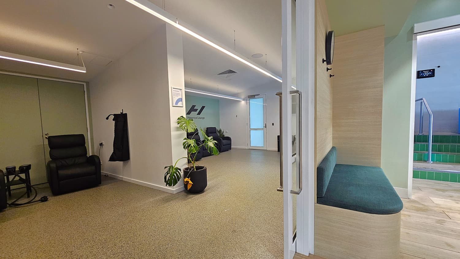On the left, a black leather recliner next to a wall with a hanging black coat. In the middle, a potted plant sits on the floor near a white column, with a sign on the wall and a door leading to another room. On the right, a built-in wooden bench with a green cushion beneath a wall-mounted clock. The floor is light brown tile, and the ceiling is white with linear recessed lighting and ventilation panels.