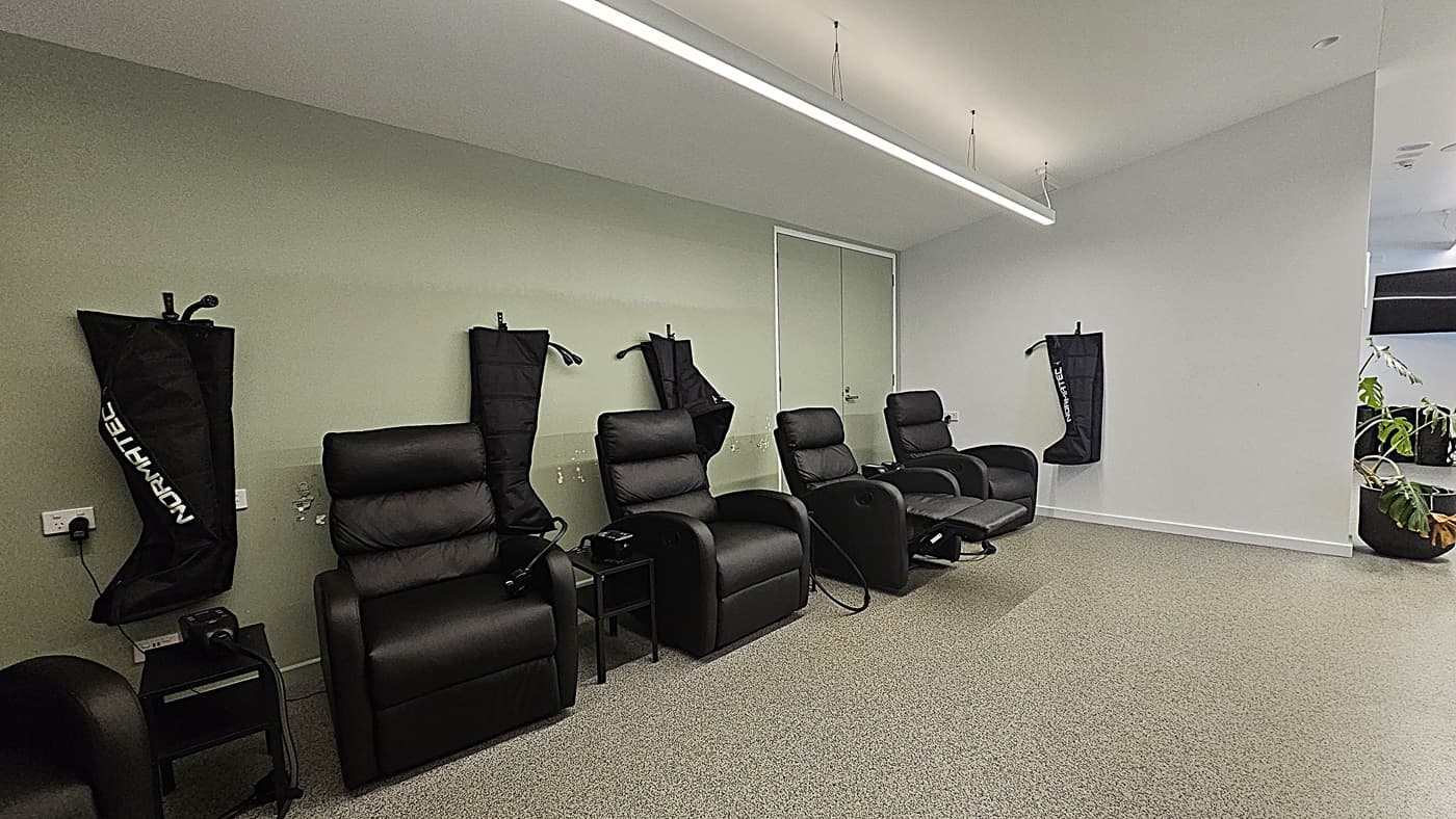 On the left, three black leather recliners with leg compression sleeves hanging on the wall behind them. In the middle, a double door with a light green wall. On the right, a single black leather recliner with a leg compression sleeve on the wall and a potted plant near the corner. The floor is grey carpet, and the ceiling is white with a long, suspended linear light fixture.
