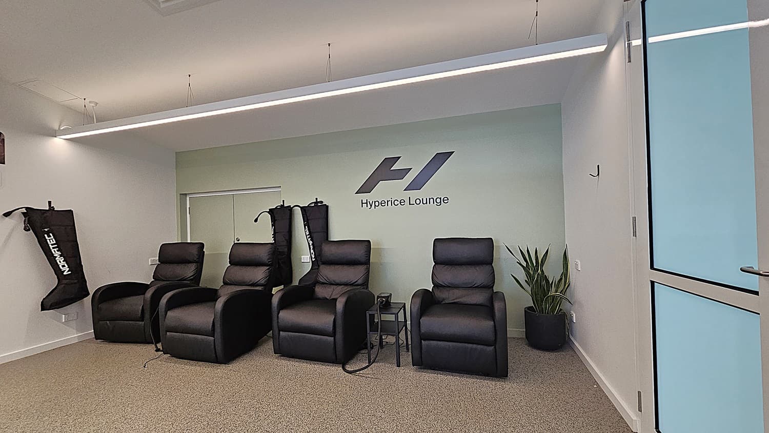 On the left, three black leather recliners with leg compression sleeves hanging on the wall behind them. In the middle, a light green wall with the "Hyperice Lounge" logo and a small side table. On the right, a potted plant next to a glass door with a metal handle. The floor is grey carpet, and the ceiling is white with a long, suspended linear light fixture.
