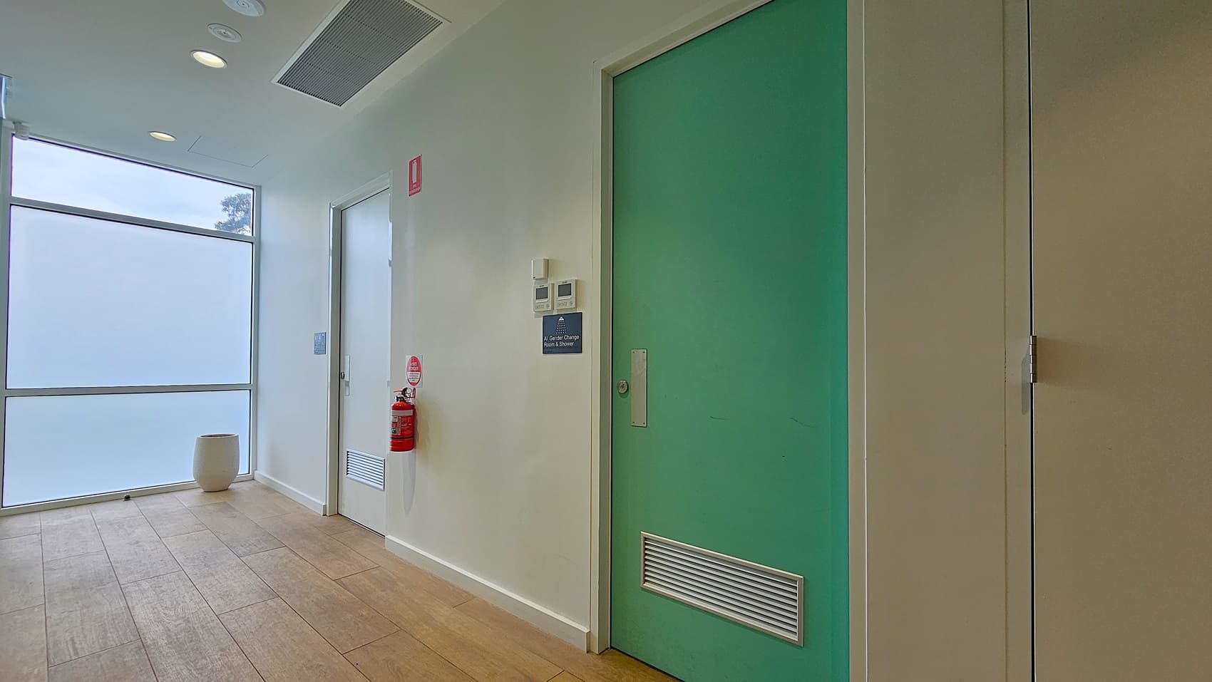 On the left, a large frosted window with a small white cylindrical stool in front of it. In the middle, a white door with a fire extinguisher mounted on the wall beside it. On the right, a green door with a ventilation grille at the bottom and a small sign above a thermostat. The floor is light wood, and the ceiling is white with recessed lighting and a ventilation panel.
