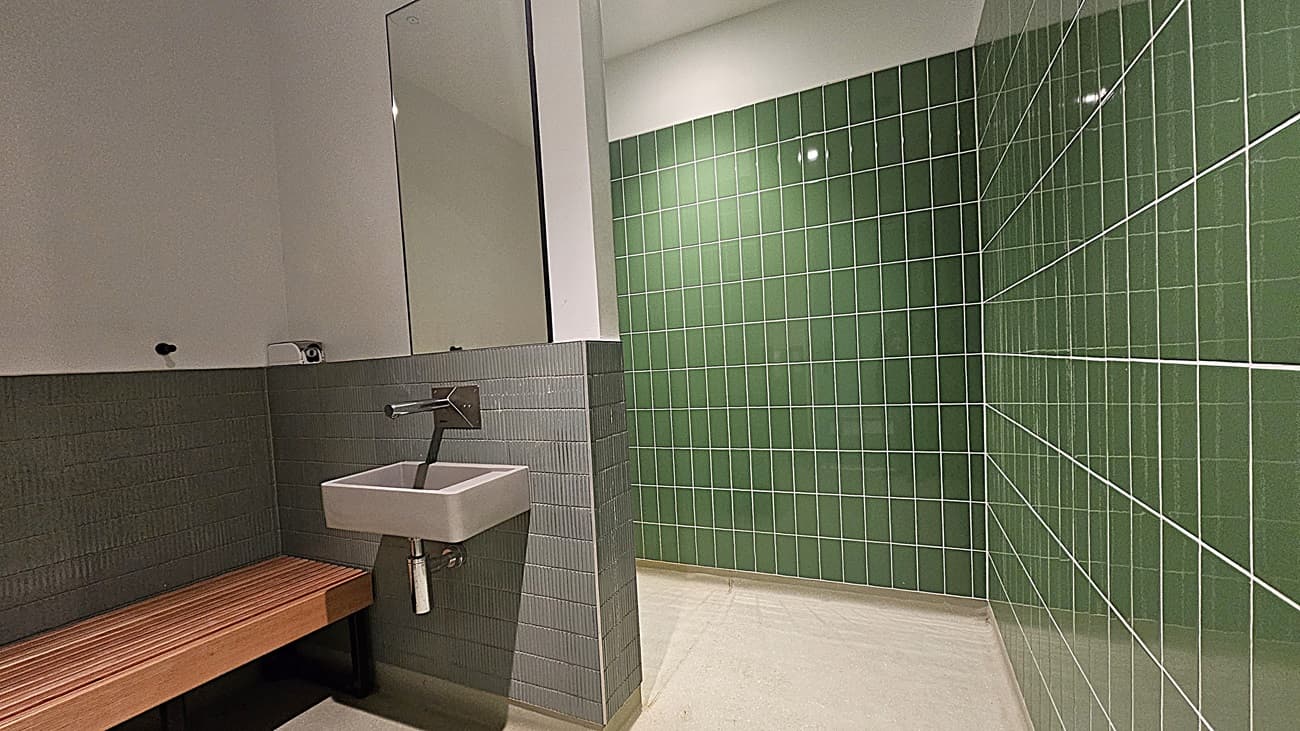 On the left, a small white sink with a wall-mounted faucet beneath a large mirror, and a wooden bench below grey tiled walls. In the middle, a white wall extends to the ceiling. On the right, green tiled walls cover the entire surface. The floor is light-coloured tile, and the ceiling is white with recessed lighting.