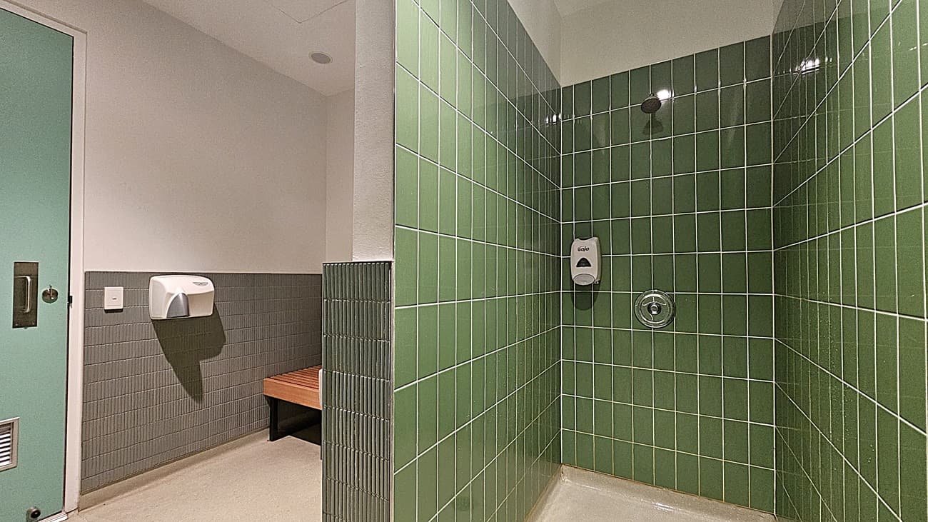 On the left, a green door with a metal handle and a wall-mounted hand dryer above grey tiles. In the middle, a wooden bench beneath grey tiles and a white wall. On the right, a shower area with green tiled walls, a soap dispenser, and a shower control knob. The floor is light-coloured tile, and the ceiling is white with recessed lighting.