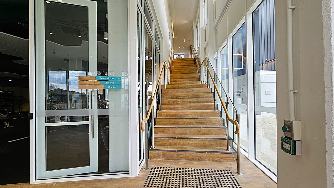 On the left, there is a glass door with a sign indicating directions to the Frawley Room, ISN Clinic, and Studios. On the right, large windows allow natural light to flood in, revealing an exterior view of a modern building. In the middle, a wooden staircase with metal railings ascends to the upper floor. The floor is wooden, and the ceiling is white with recessed lighting.