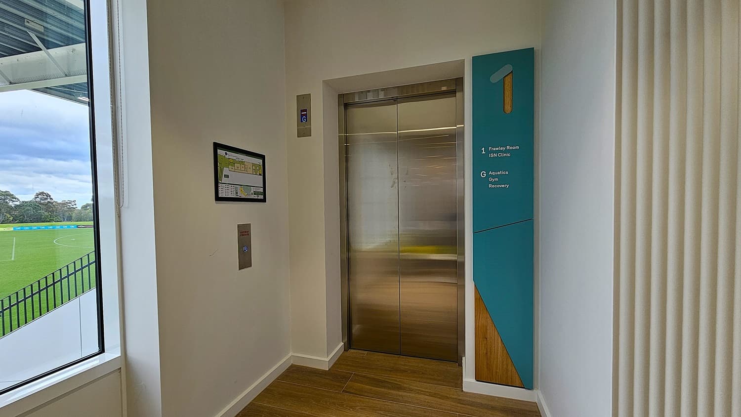 On the left, a large window reveals a green sports field with a black railing. In the middle, a stainless steel lift with closed doors is flanked by a control panel and a wall-mounted map. On the right, a blue sign indicates directions to the Frawley Room, ISN Clinic, Aquatics, Gym, and Recovery. The floor is wooden, and the ceiling is white.