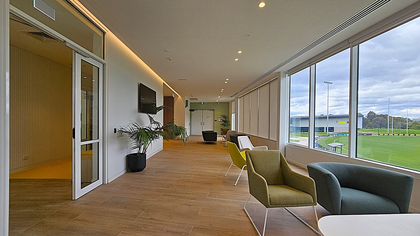 On the left, a glass door leads to another room, and a potted plant sits beside a wall-mounted television. In the middle, a corridor with wooden flooring extends towards double doors at the far end, flanked by more potted plants. On the right, large windows with roller blinds provide a view of a green sports field and an adjacent building. The ceiling is white with recessed lighting.