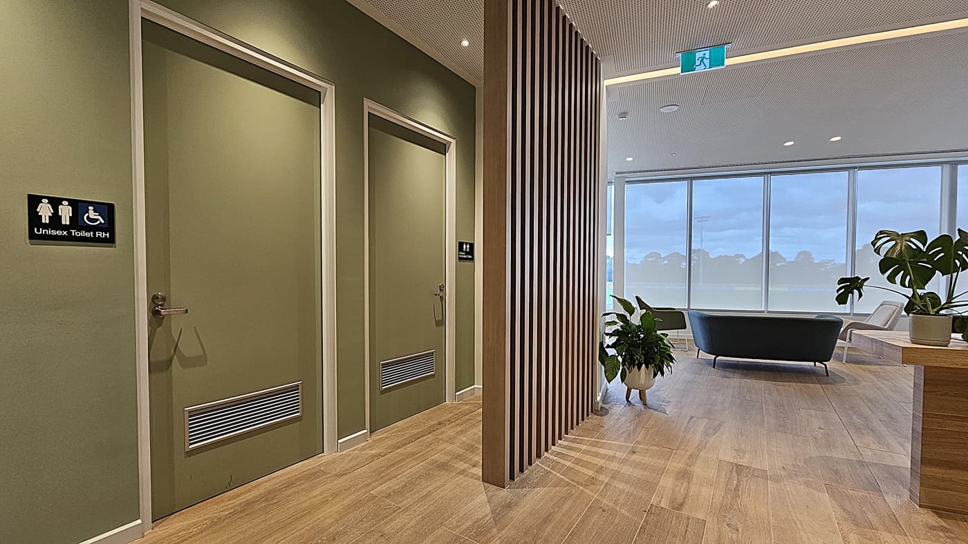 On the left, there are two green doors with signs indicating unisex toilets, one of which is accessible. In the middle, a wooden slatted partition partially divides the space. On the right, large windows provide a view of an outdoor area, and a seating area with a black sofa and potted plants is visible. The floor is wooden, and the ceiling is white with recessed lighting and an emergency exit sign.