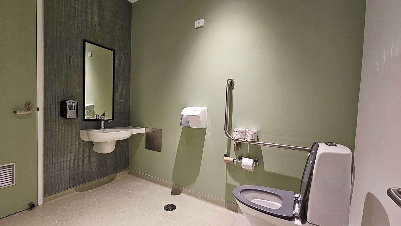 On the left, a green door with a handle and a ventilation grille is next to a wall-mounted soap dispenser and a rectangular mirror above a white sink. In the middle, a paper towel dispenser is mounted on the green wall. On the right, a white toilet with a grey seat is equipped with a grab rail, toilet paper holder, and two rolls of toilet paper on a shelf. The floor is light-coloured and textured, and the ceiling is white with a small light fixture.
