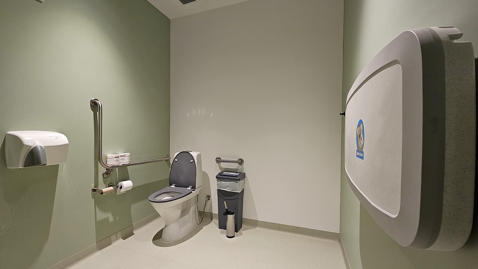 On the left, a wall-mounted paper towel dispenser is next to a grab rail and a white toilet with a grey seat. In the middle, a small grey bin and a toilet brush are placed against the wall. On the right, a baby changing station is mounted on the wall. The floor is light-coloured and smooth, and the ceiling is white with a small light fixture.
