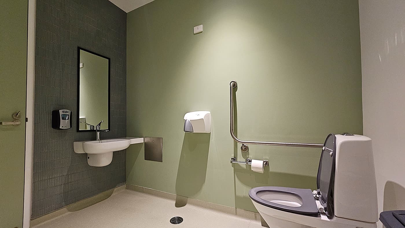 On the left, a white sink with a wall-mounted soap dispenser is below a rectangular mirror on a tiled wall. In the middle, a paper towel dispenser is mounted on a green wall. On the right, a white toilet with a grey seat is equipped with a grab rail and a toilet paper holder. The floor is light-coloured and smooth, and the ceiling is white with a small light fixture.